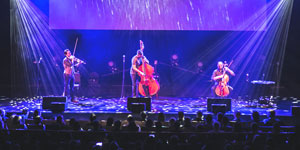 Three musicians perform on stage under blue lights; two play violins and one plays a cello, with an artistic background projection enhancing the ambiance.