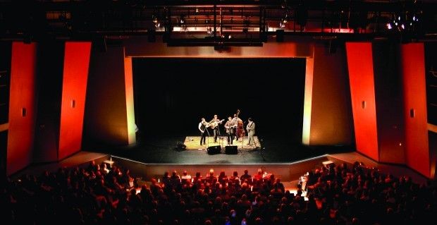 A band performing on a well-lit stage in a theater with orange walls and an audience in dark seating watching attentively.