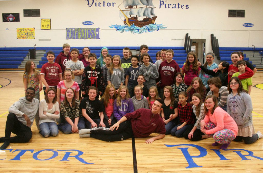 5695bf2cbb6fe.image A large group of diverse students posing for a photo in a gymnasium with the words "victor pirates" on the wall behind them. some students are sitting, others standing, all smiling at the camera.