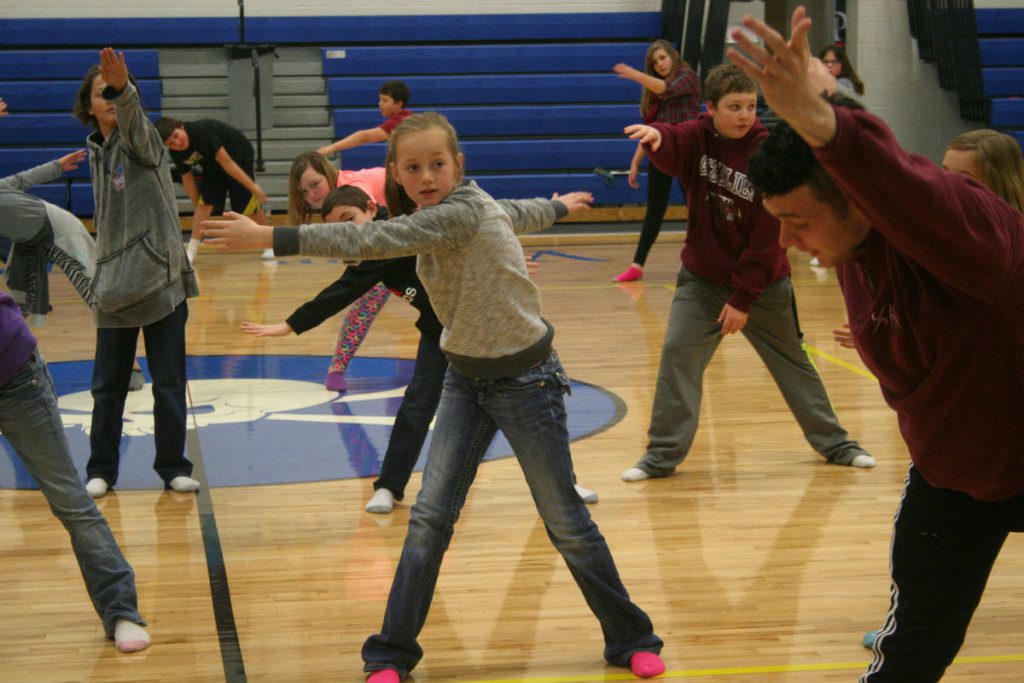 5695c01e0a513.image A group of children and a young adult in a gymnasium are spread out, engaging in a dance or exercise routine with arms extended and concentrating on their movements.