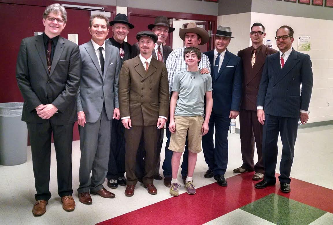 bbvdgroup Group of nine men and one boy posing in a hallway, dressed in various styles of suits and hats, ranging from modern to vintage. they stand smiling, against a backdrop of red lockers.