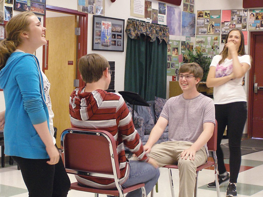 rsc_workshop3 Four teenagers engage in a lively conversation in a school hallway decorated with photographs and posters. two are seated on chairs, while the other two stand, all smiling and gesturing expressively.