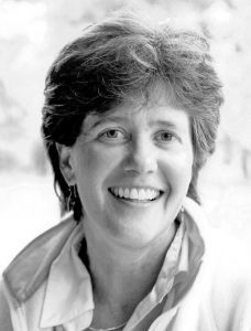 Laura-Merrill Black and white portrait of a smiling middle-aged woman with short, curly hair, wearing a jacket and a collared shirt. she appears cheerful against a blurred background.