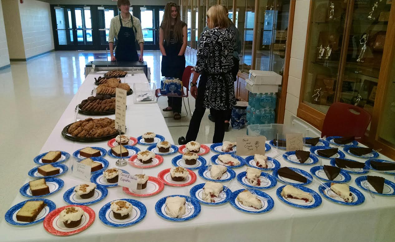 bbvdrefresh A school bake sale with a variety of cookies and pies displayed on a table. two people stand talking, another person walks past.