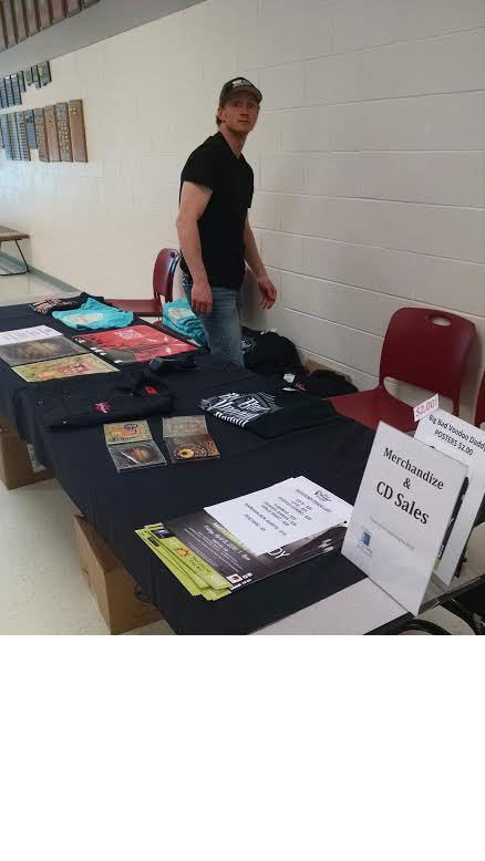 merch A young man stands behind a table displaying various merchandise like cds and t-shirts at an indoor event, with a sign titled "merchandise cd sales" next to him.