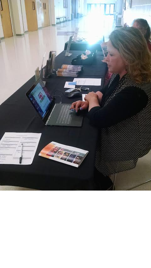 volunteer1 A woman concentrates on her laptop at a registration table with forms and informational brochures, in a brightly lit hall.