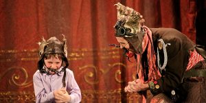 A child wearing a medieval helmet and a man dressed in a quirky steampunk costume interact cheerfully on a stage with a lavish red curtain backdrop.
