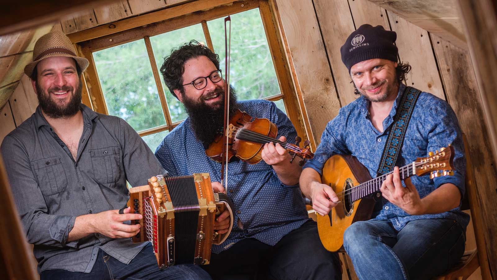 Three smiling men playing folk music inside a cozy wooden cabin, one with a guitar, one with a violin, and one with an accordion.