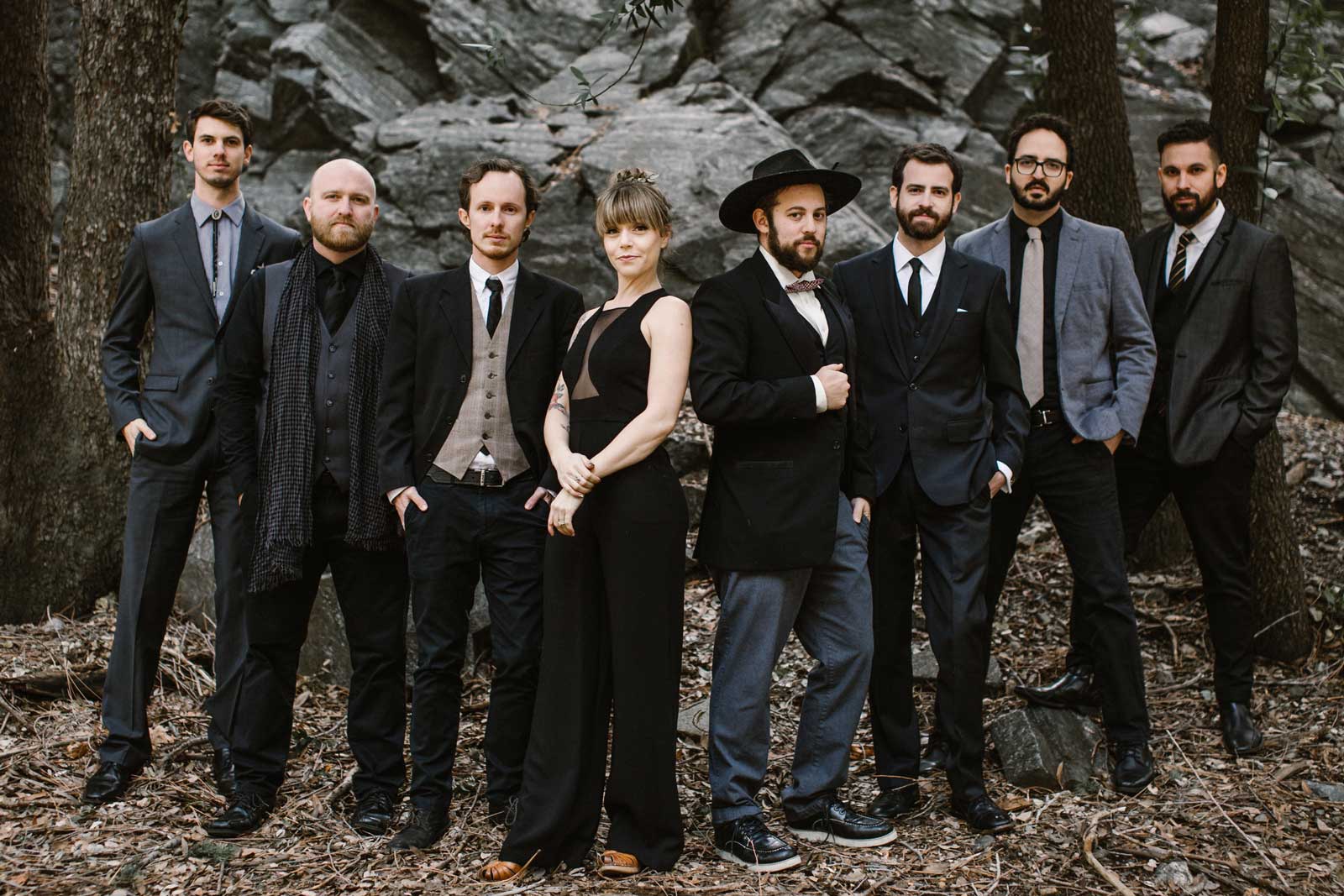 A group of eight people, dressed in formal attire, stand together in a wooded area. the group, consisting of seven men and one woman, exhibit a variety of styles, from suits to a hat.
