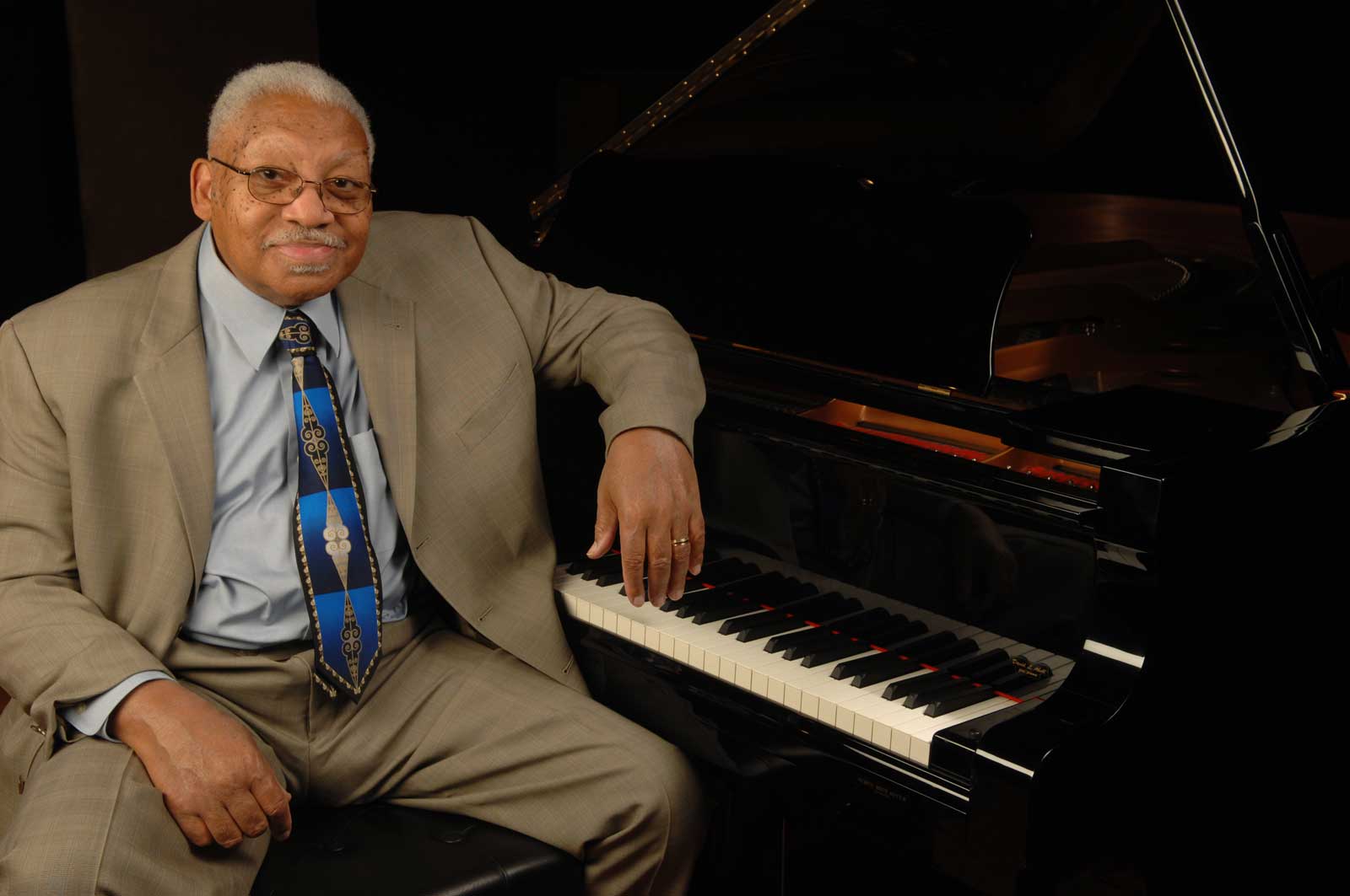 An elderly man with glasses, wearing a beige suit and patterned tie, sits beside a grand piano, smiling gently with one hand on the piano keys in a dimly lit room.