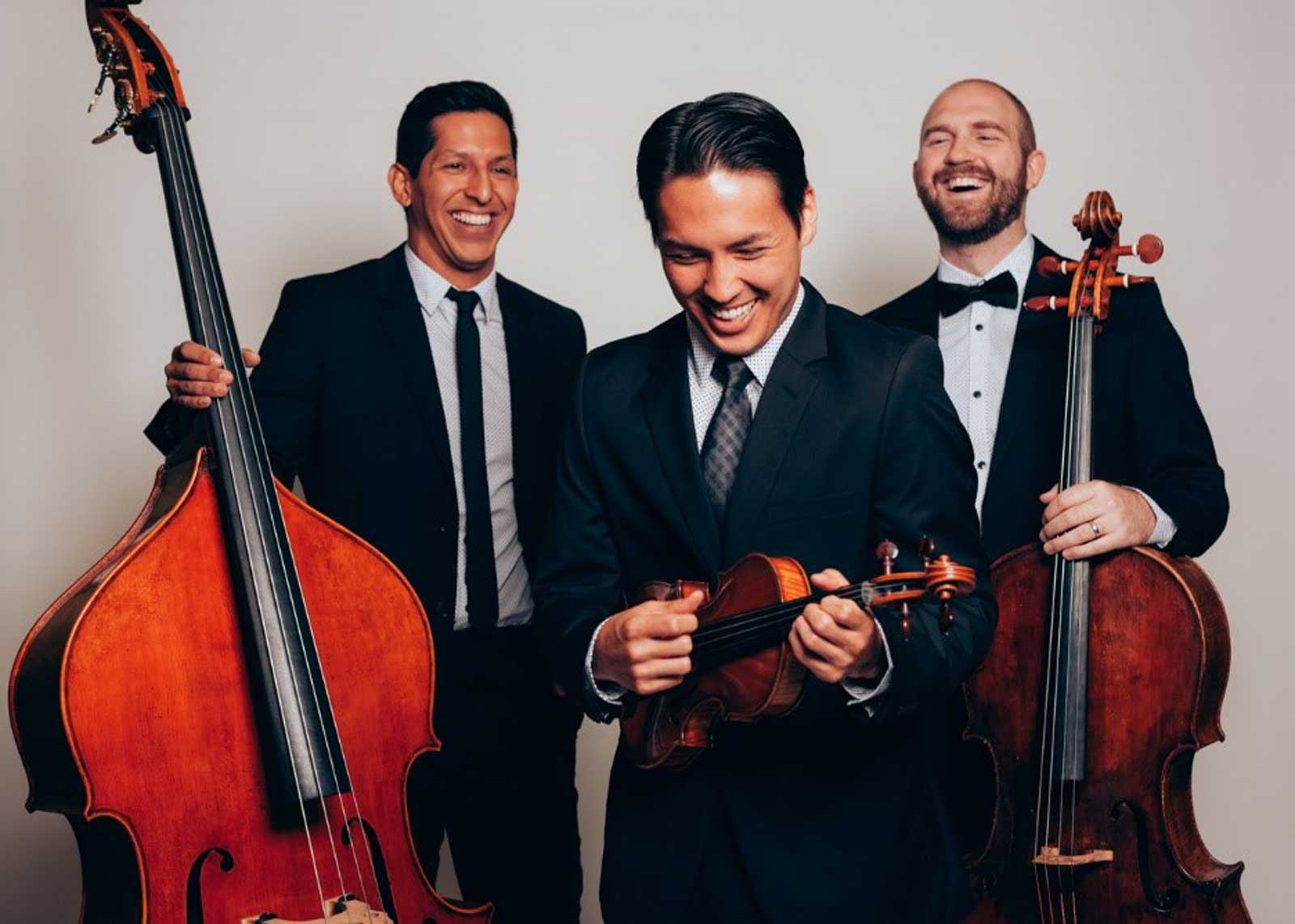 Three smiling men in formal wear, one playing a violin and two holding upright basses, stand against a light gray background, sharing a joyful moment.