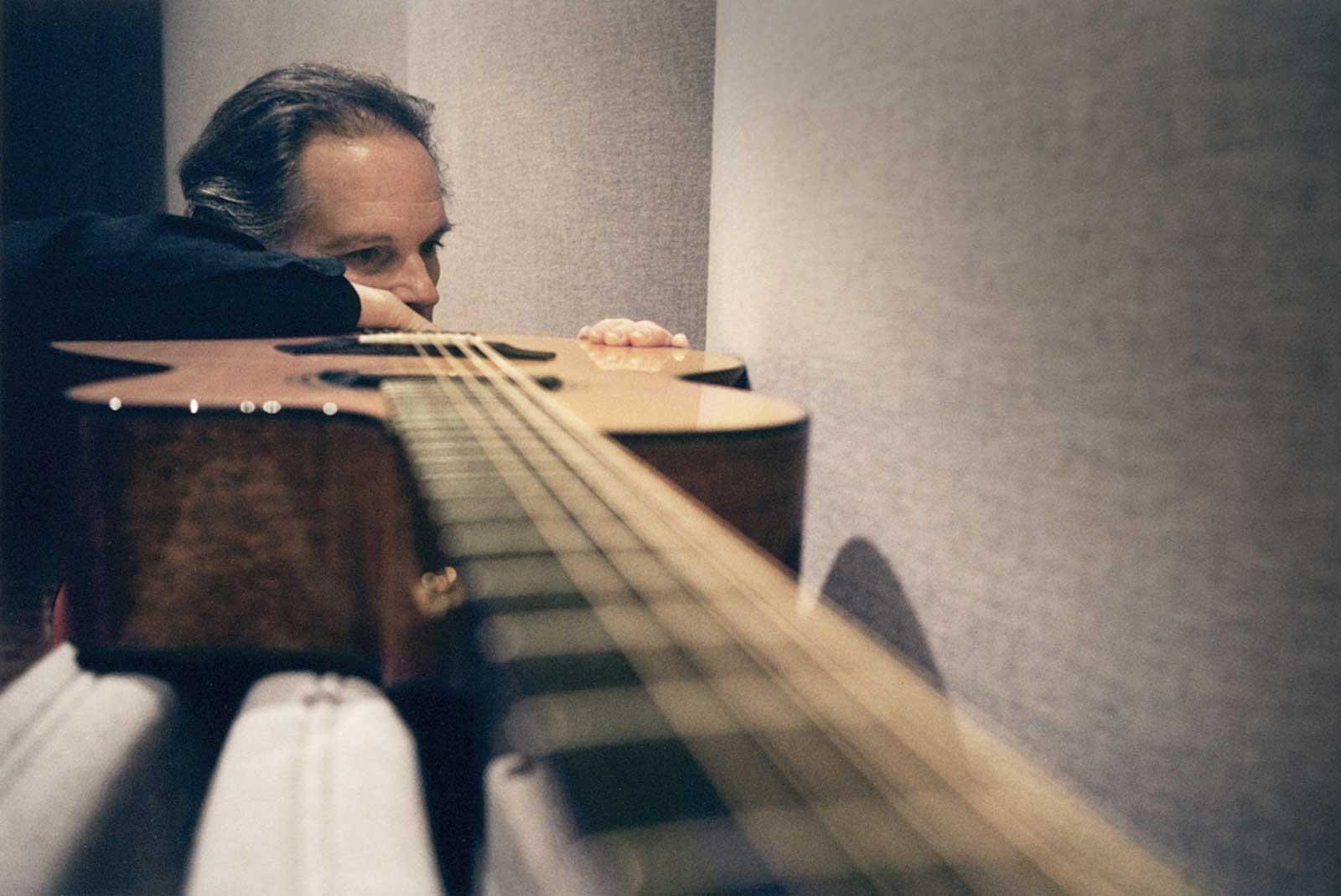 A man rests his head on his arm, gazing thoughtfully along the fretboard of an acoustic guitar, with focus on the strings and instrument while the background is softly blurred.