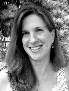 Kristina-Bessenyey A black and white portrait of a smiling woman with shoulder-length hair, wearing earrings, standing in front of softly focused pine branches.