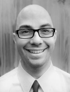 chris-web Black and white portrait of a smiling bald man wearing glasses and a white shirt, standing in front of a wooden panel background.