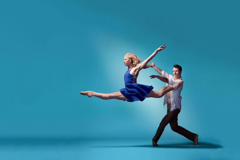 A female ballet dancer in a blue dress performs a graceful leap, supported by a male dancer in brown trousers and a white shirt against a light blue background.
