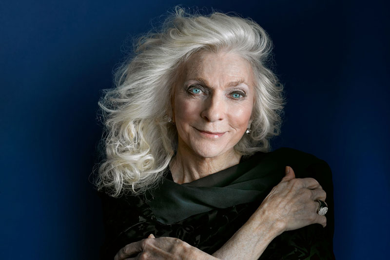 An elderly woman with long, wavy gray hair and a warm smile, wearing a black outfit, against a deep blue background. her hands are gracefully positioned on her shoulder.