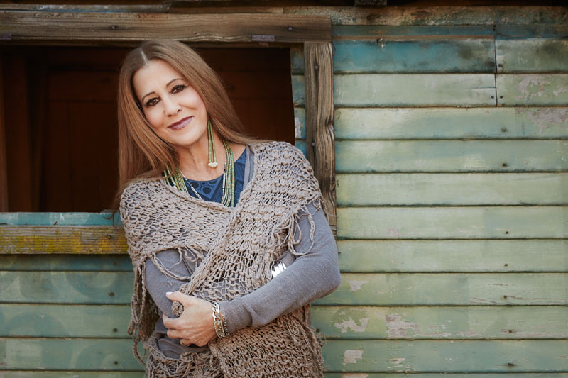 A smiling woman leaning on a wooden window frame of a rustic, blue-green shack. she is wrapped in a shawl, wearing a gray top and layered necklaces.
