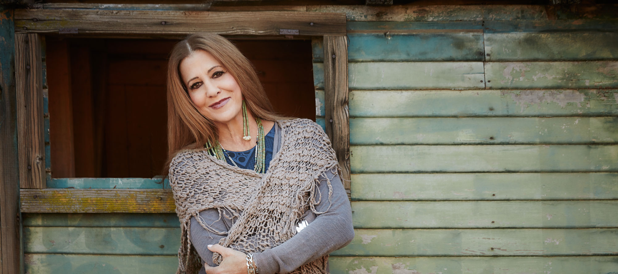 A woman with a gentle smile leans against a weathered wooden shack, wearing a grey knitted shawl over a blue top, exuding a relaxed and confident pose in a rustic setting.