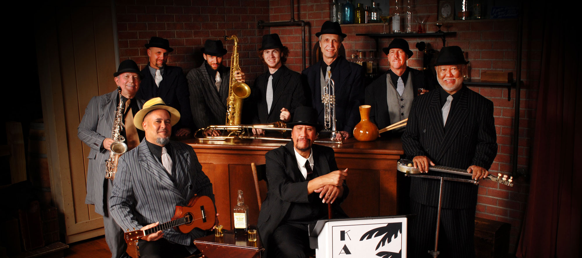 A jazz band of nine men dressed in suits and fedoras posing with their instruments, including saxophones, a guitar, and a double bass, in a dimly lit room.