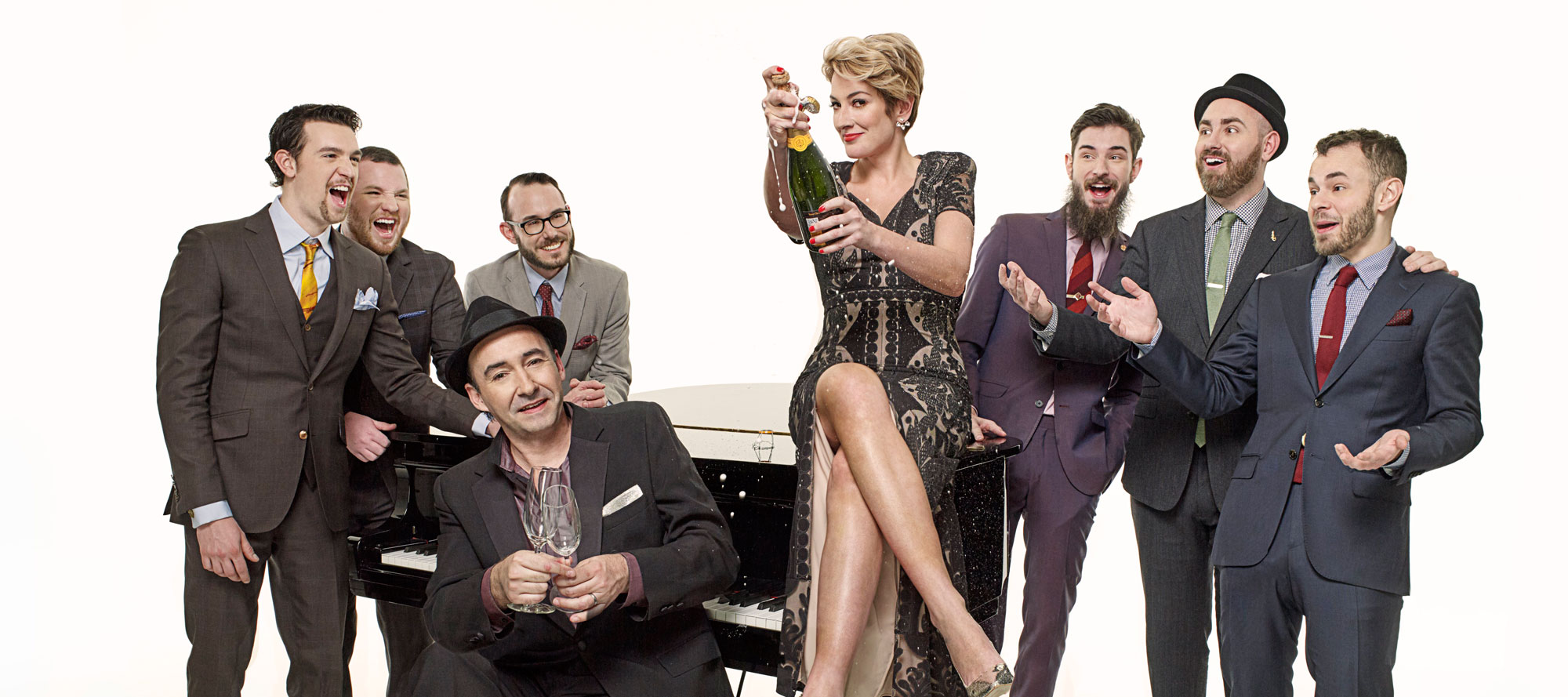 Group of six men and one woman, all smartly dressed, around a black piano. one man plays piano, others cheer, one woman holding a glass, all smiling, on a white background.