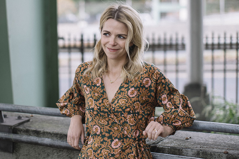 A smiling woman in a floral dress leans on a concrete railing, with a blurred background of green railings and trees.