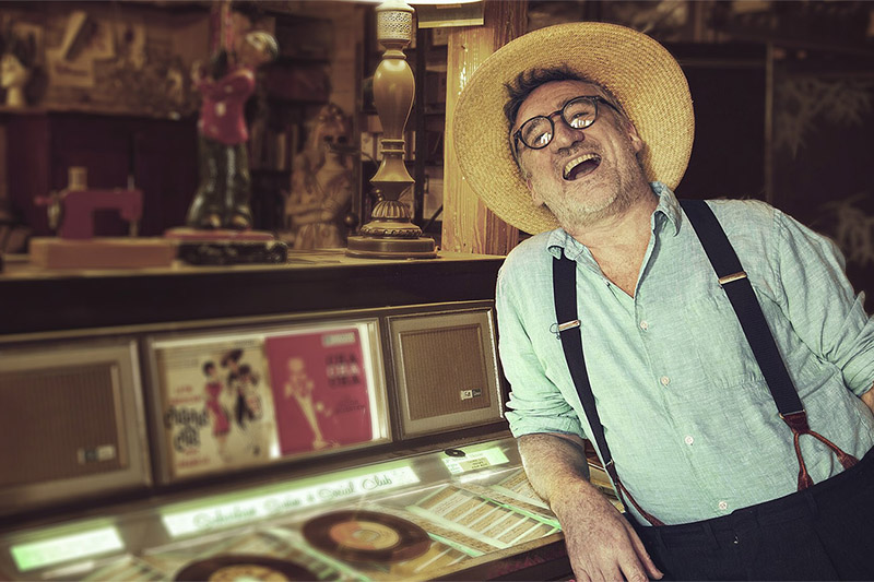 Joyful man wearing a straw hat and suspenders laughs while leaning on a vintage jukebox in a room filled with nostalgic memorabilia.