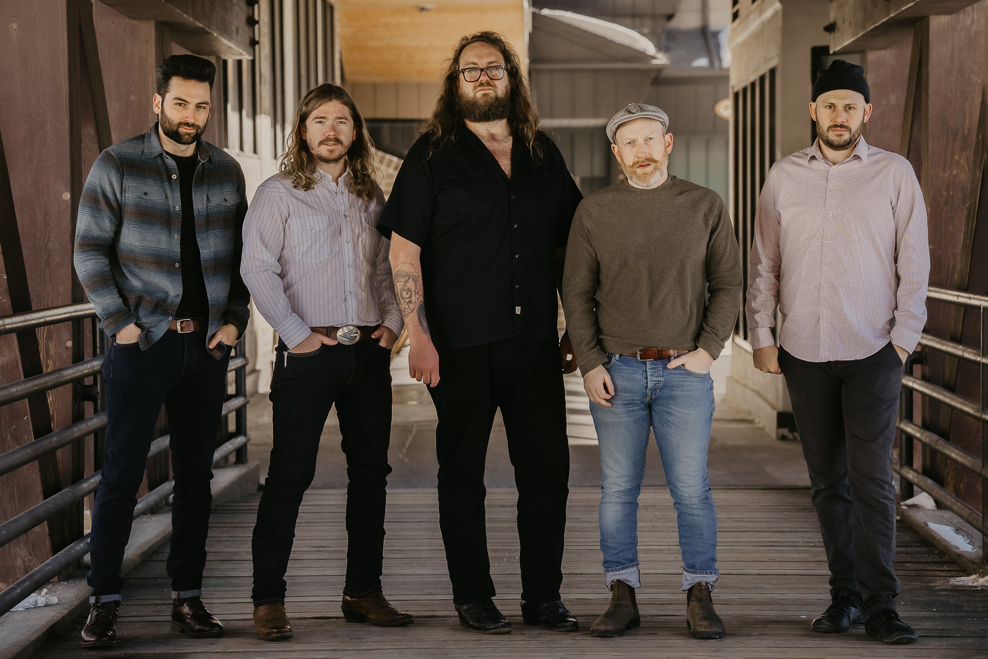 Five men stand confidently on a wooden walkway, dressed in casual and semi-formal attire, projecting a cohesive and stylish group appearance.