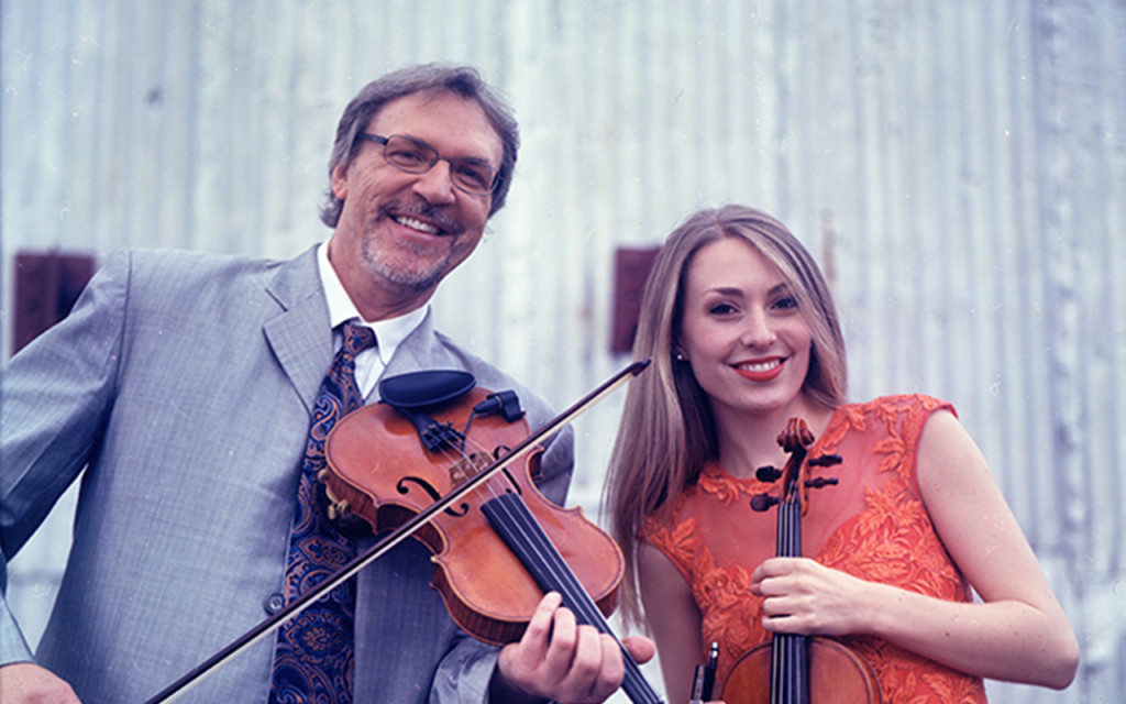 Two smiling violinists, a man and a woman, standing in front of a corrugated metal background, holding their violins. the man is wearing a suit and tie, and the woman is in an orange dress.