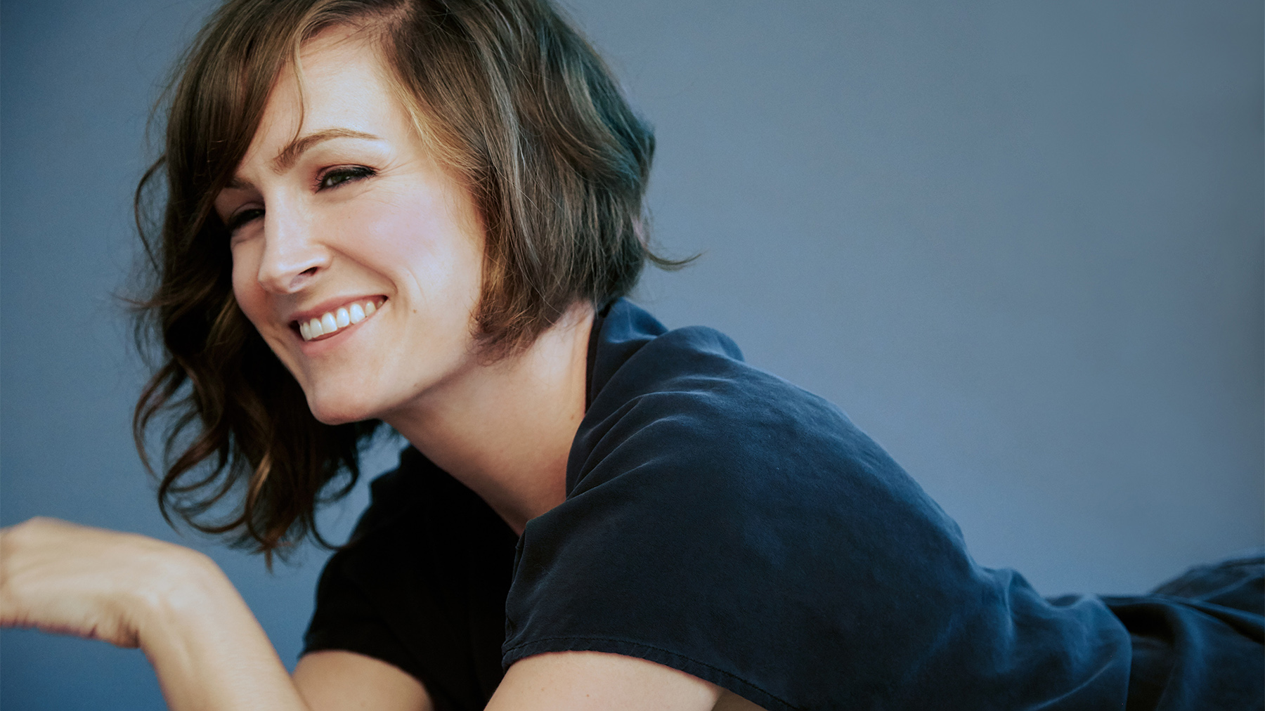 A joyful woman with short brown hair, wearing a dark blue t-shirt, smiles warmly while lying on her side against a grey background.
