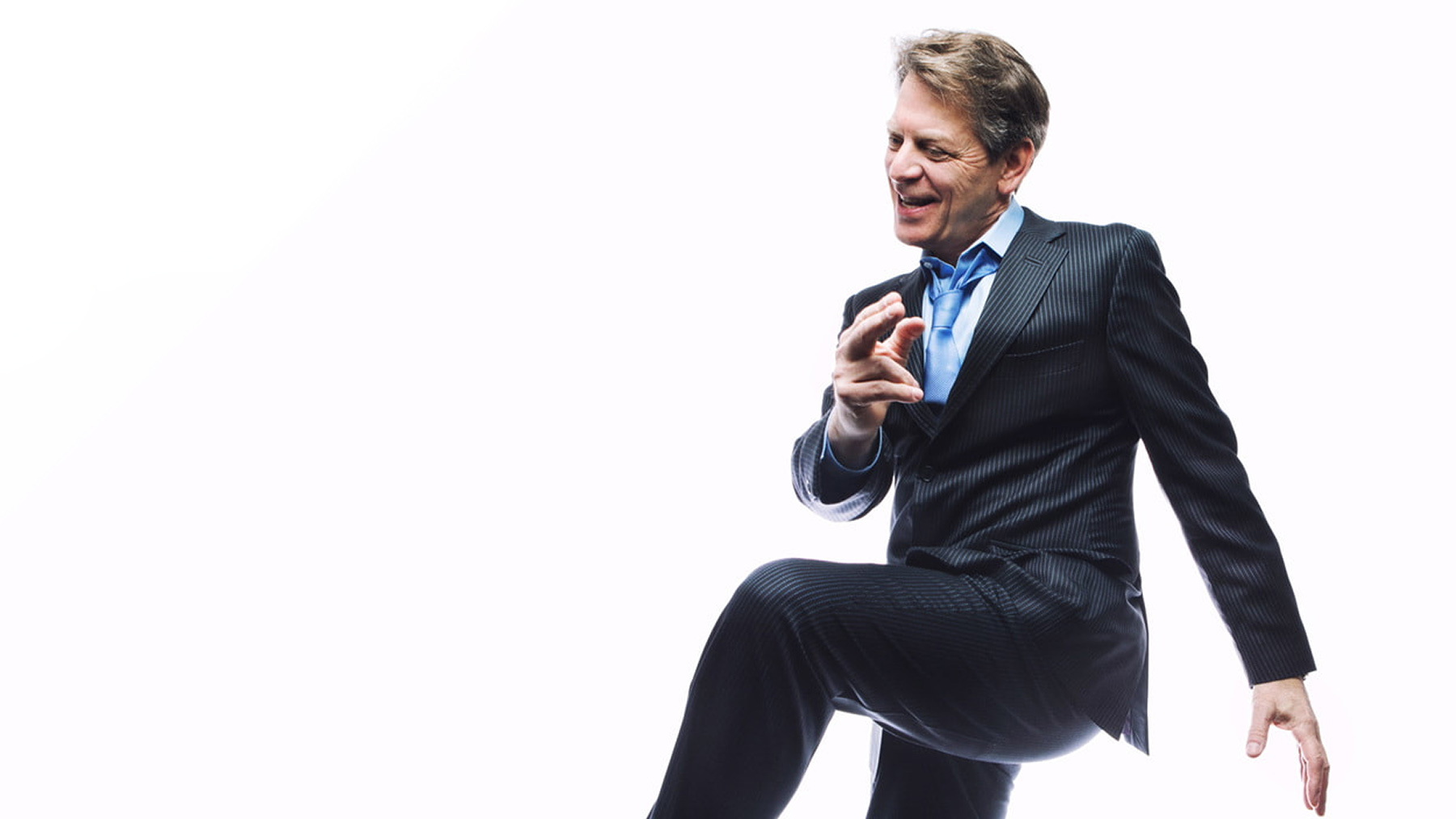 A man in a business suit playfully lifts his knee, pretending to climb an invisible step, while smiling and looking forward, isolated on a white background.