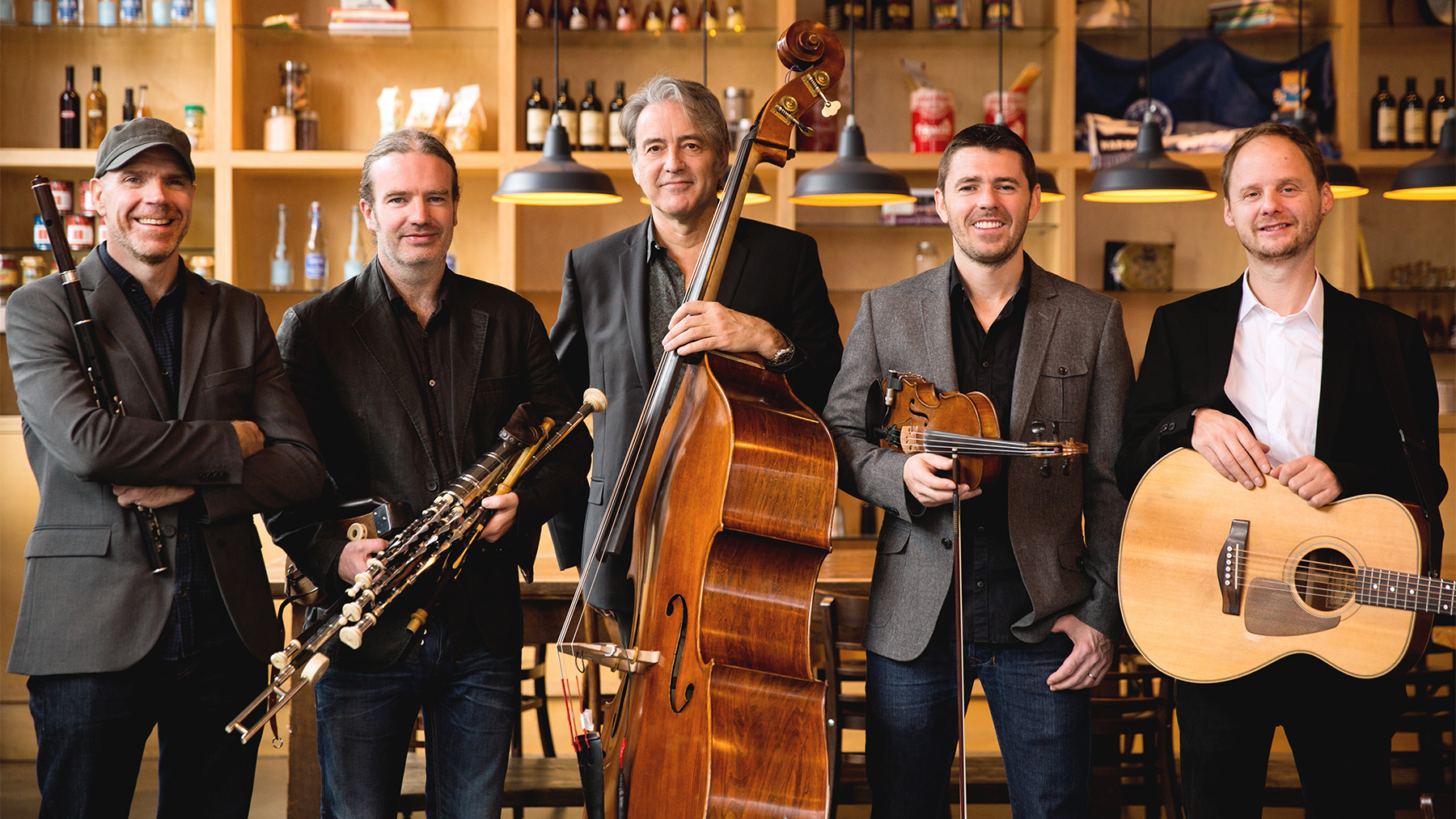 Five men holding musical instruments (clarinet, double bass, violin, guitar) smiling in a cozy bar with shelves of bottles in the background.