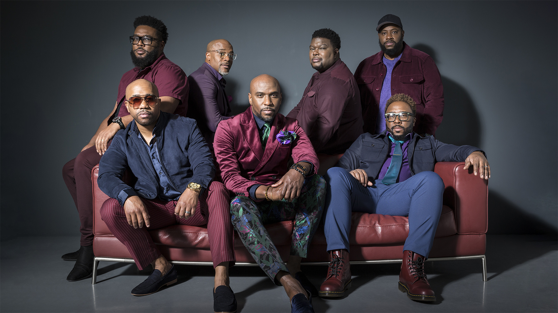 Seven stylish men in fashionable attire seated and standing around a luxurious sofa against a dark backdrop. they exhibit confident poses and expressions.