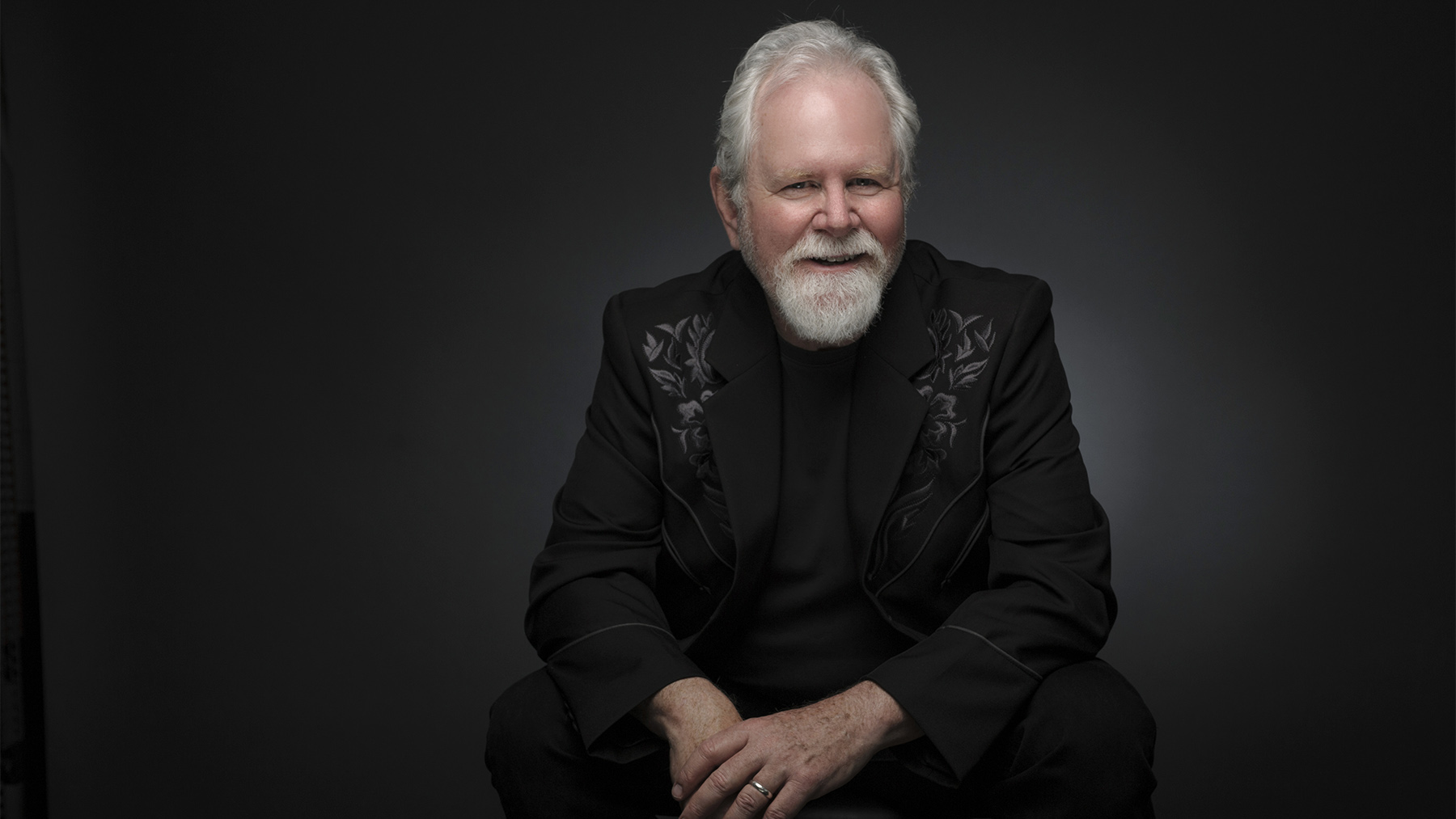 An older man with a white beard smiles gently while seated, wearing a black suit with embroidered details. he is set against a dark grey background.