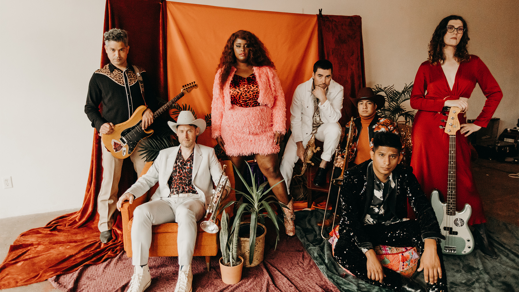 A band of seven diverse members posing with musical instruments in a room with earthy toned decor, featuring eclectic clothing styles ranging from a white suit to a vibrant pink outfit.