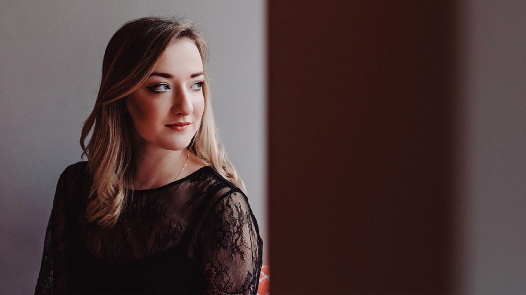 A woman in a black lace dress looks thoughtfully to the side, her blonde hair styled softly. the ambient lighting casts a warm glow on her face.