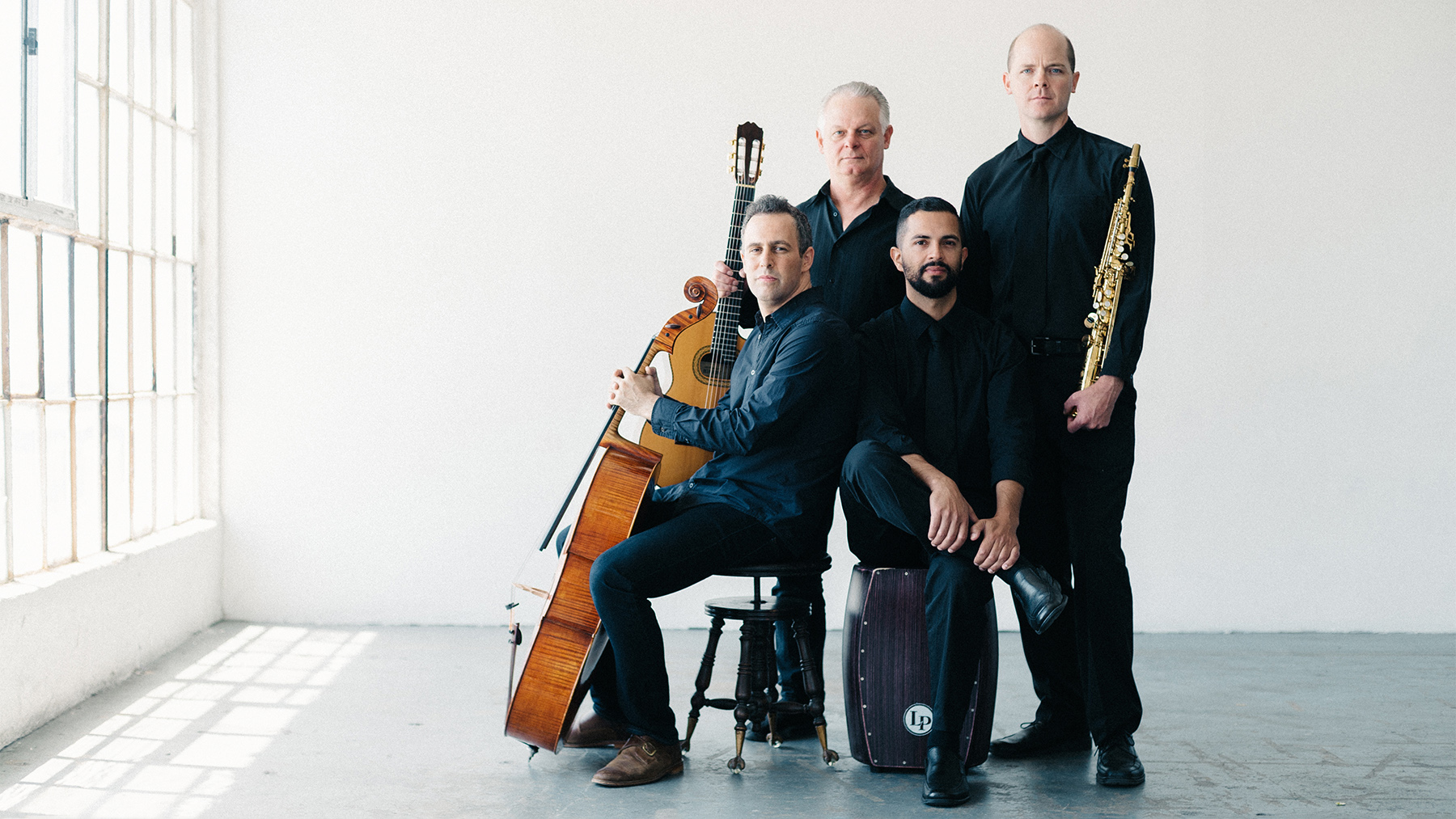 Four male musicians pose with their instruments in a bright room. two are standing, two seated, holding a cello, a double bass, and two sit with percussion instruments.