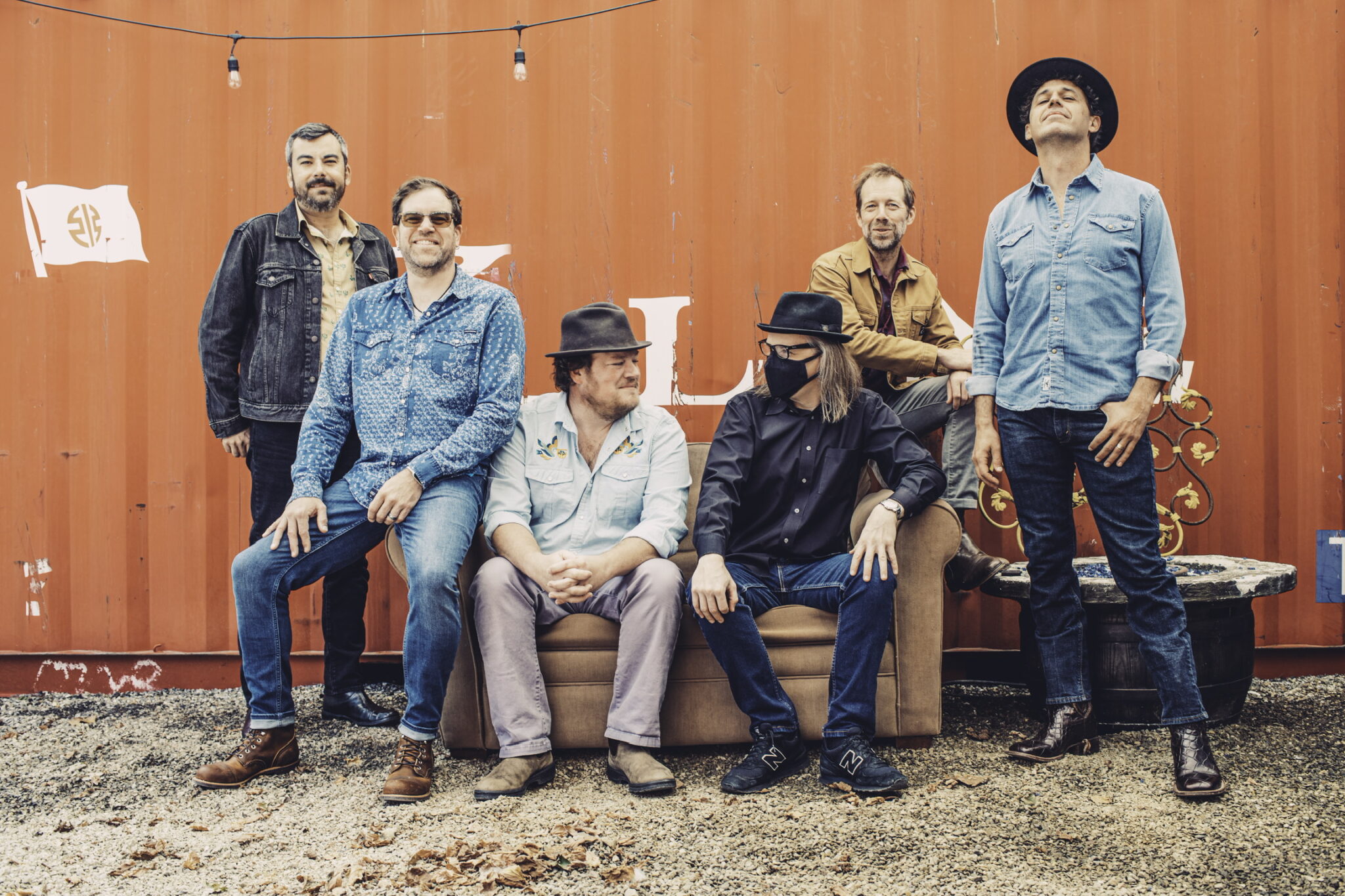 A band of six men posing casually in front of a red shipping container, dressed in denim and hats, exuding a relaxed, cool vibe.