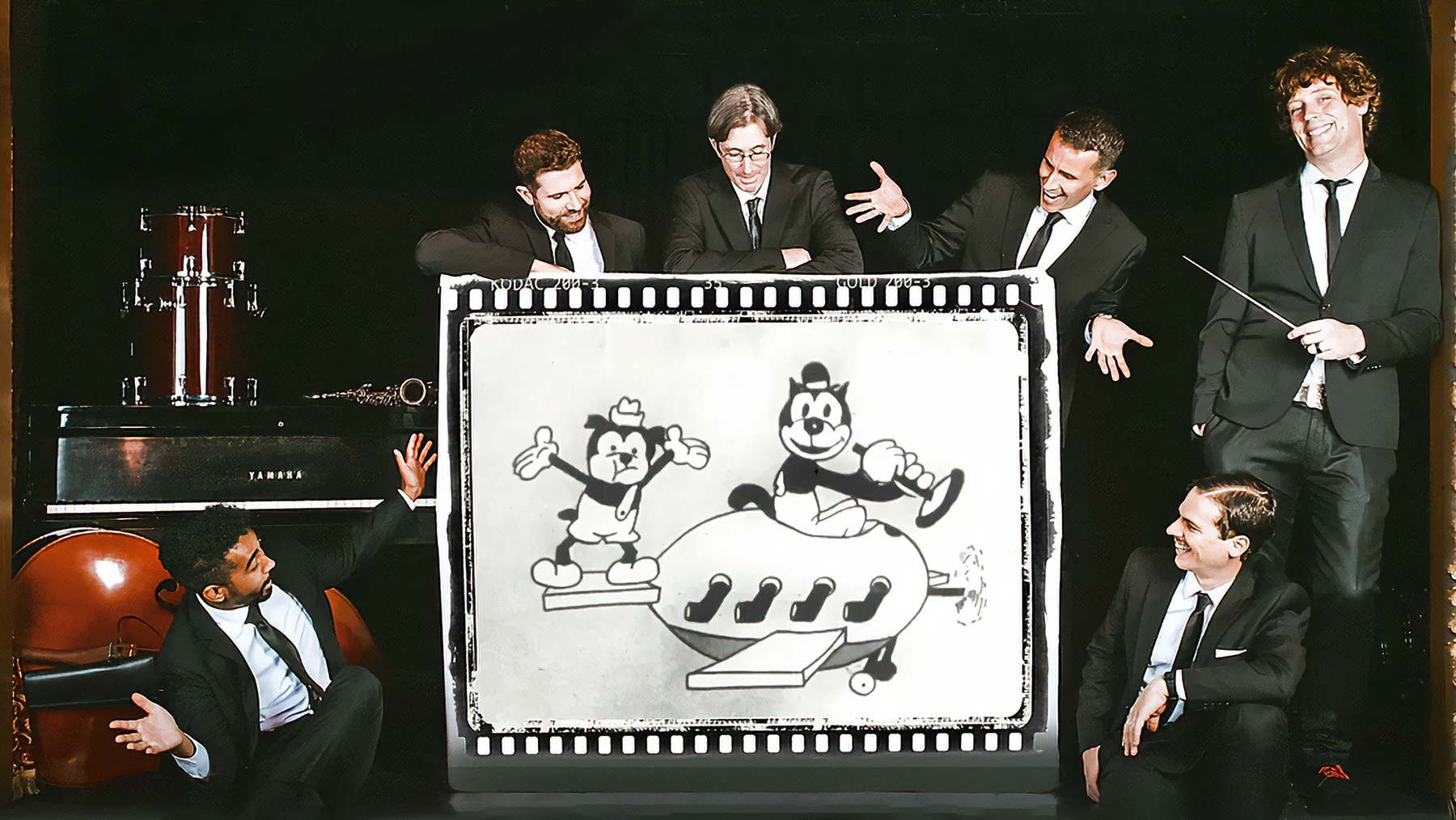 Five men in suits interact playfully around a retro cartoon projection featuring animated characters. the setting includes a piano and instruments, creating a whimsical, theatrical atmosphere.