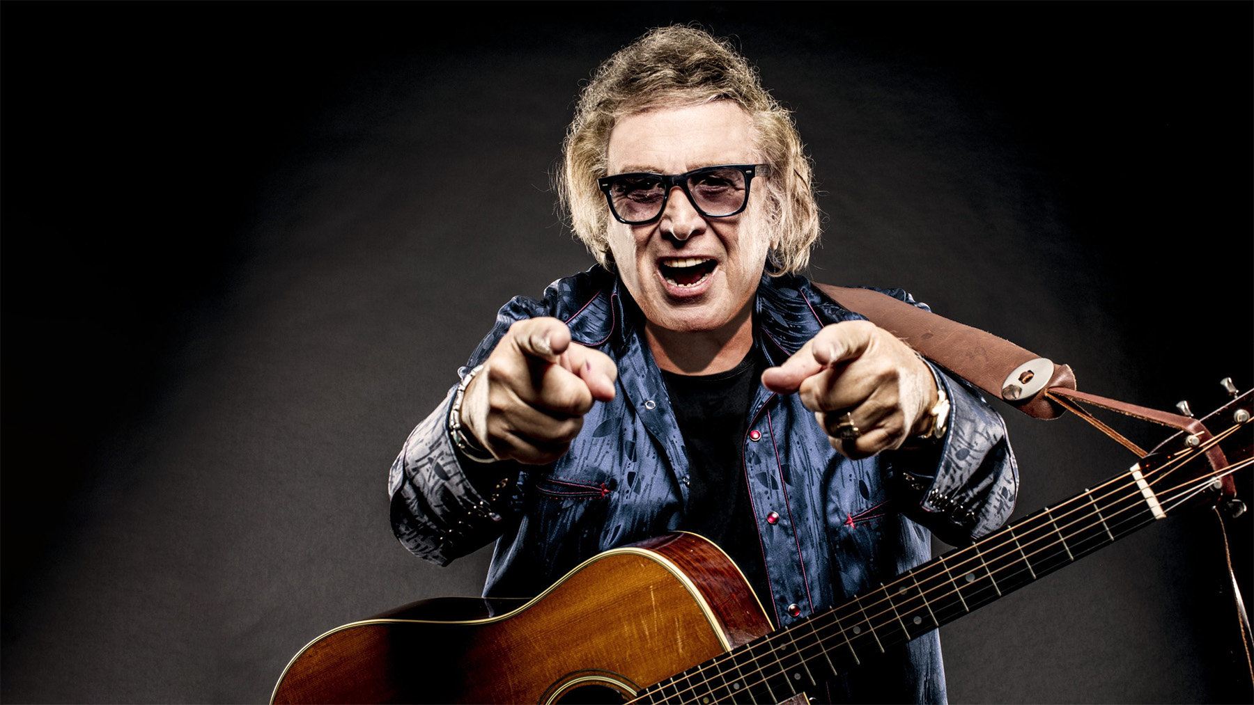 Rock musician with tousled hair wearing sunglasses and a denim jacket, pointing forward, holding an acoustic guitar, set against a dark background.