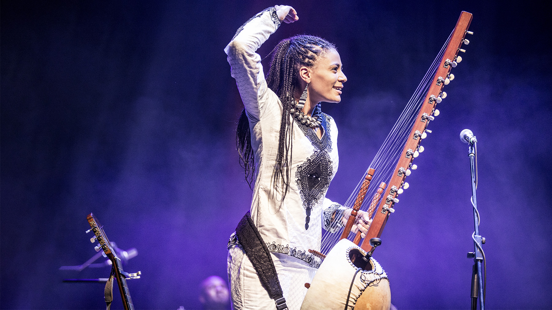 A female musician stands on stage, triumphantly raising her fist while playing a large, traditional stringed instrument, dressed in an ornate white and black outfit, under vibrant stage lighting.