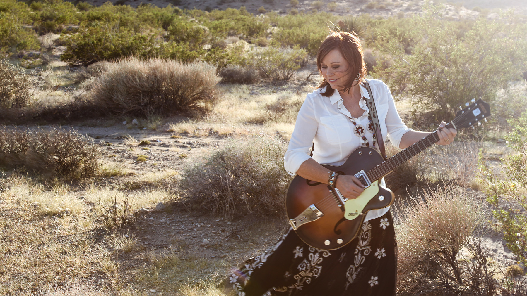 A woman plays an acoustic guitar in a desert setting, with sunlight highlighting her and the surrounding bushes. she is dressed in a white blouse and patterned skirt, focused on her instrument.