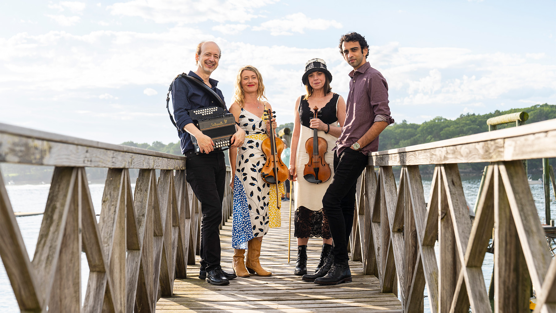 Four musicians on a wooden pier overlooking a lake, with the man on the left holding an accordion, two women in the center (one with a cello, the other with a violin), and a man on the right with a guitar.