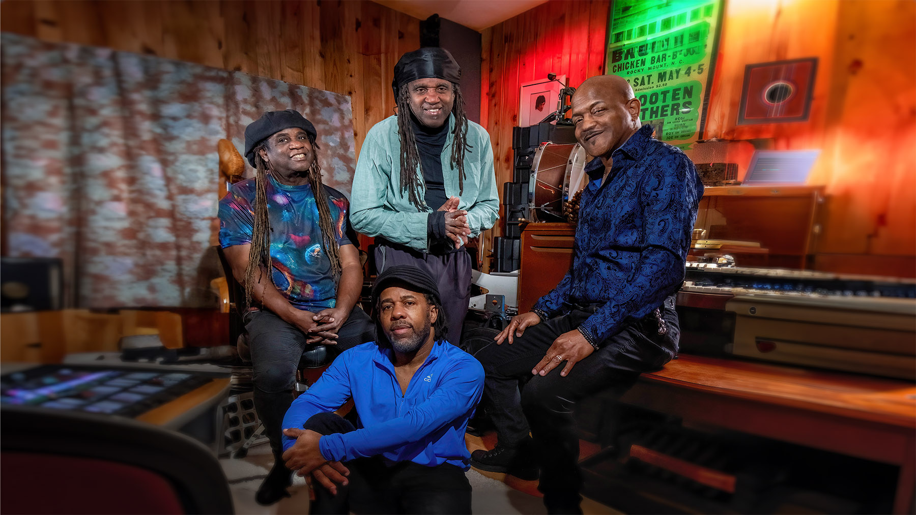 Four smiling men in a warmly lit music studio. they sport colorful attire and are surrounded by musical instruments, including keyboards and a guitar.