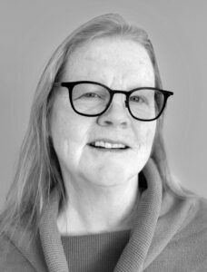 JABioPic copy A black and white portrait of an older woman with long gray hair, wearing glasses, and a cowl neck sweater, smiling gently at the camera.