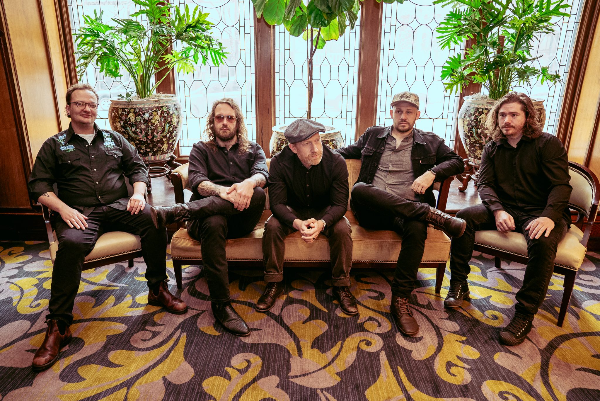 Five men sit on a settee in a stylish room with large windows and plants. They all wear dark clothing, ranging from casual to slightly formal. Their expressions are serious, creating a contemplative atmosphere. The room has a patterned carpet and intricate window designs.
