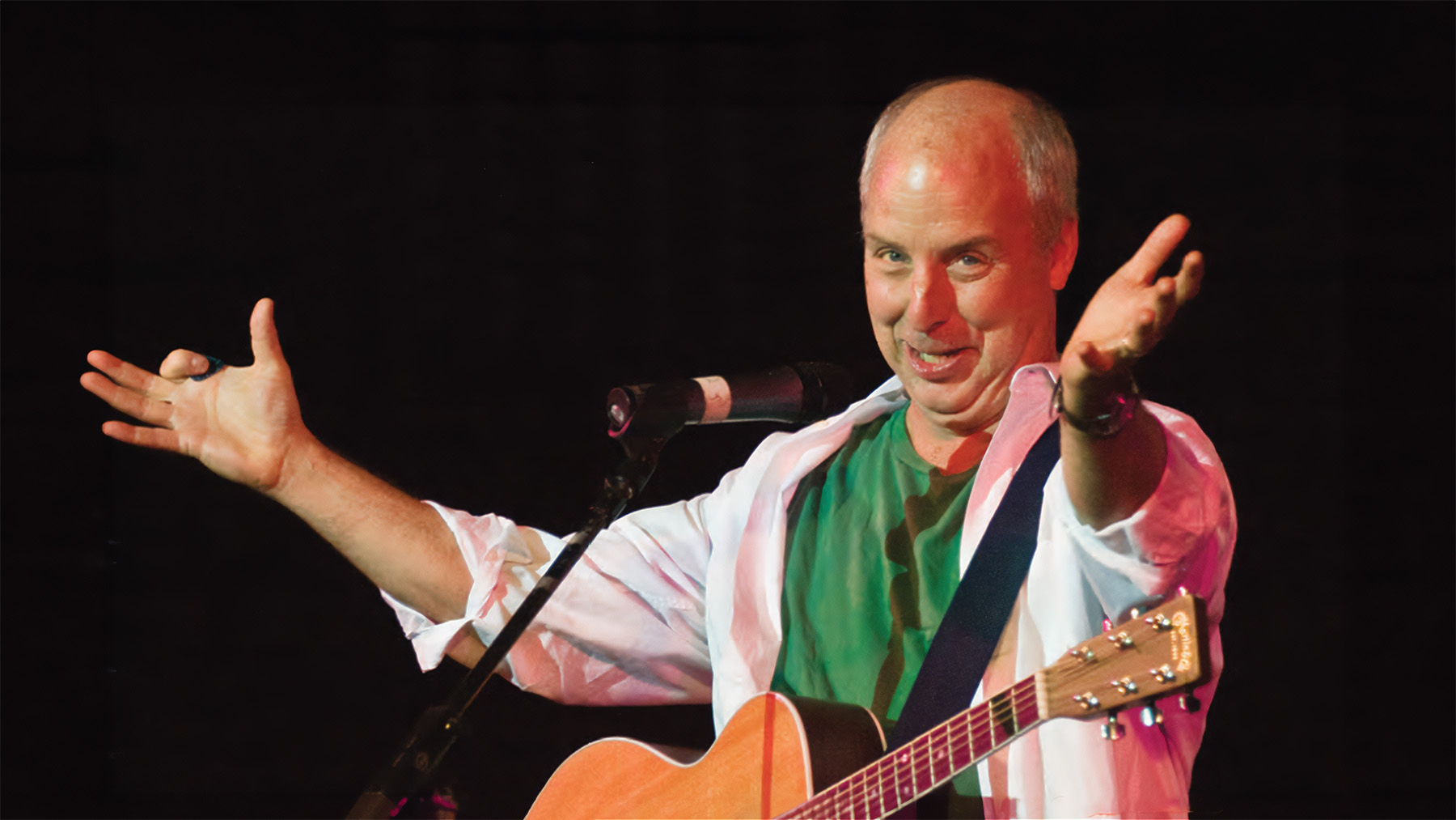 A male singer with a guitar on stage, enthusiastically talking into a microphone with his hands spread wide, wearing a white and pink shirt. he has a joyful expression.