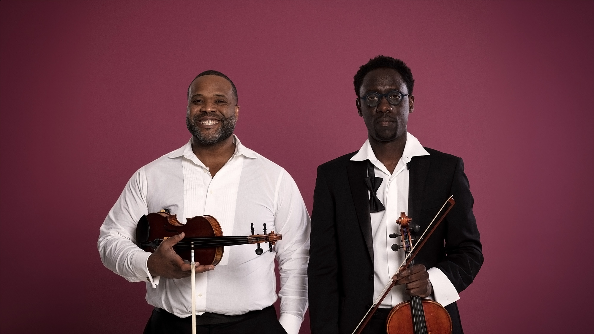 Two men holding violins pose against a maroon background. The man on the left, in a white shirt, smiles, while the man on the right, wearing glasses and a black suit, looks serious. Both have their violins positioned in front of them.