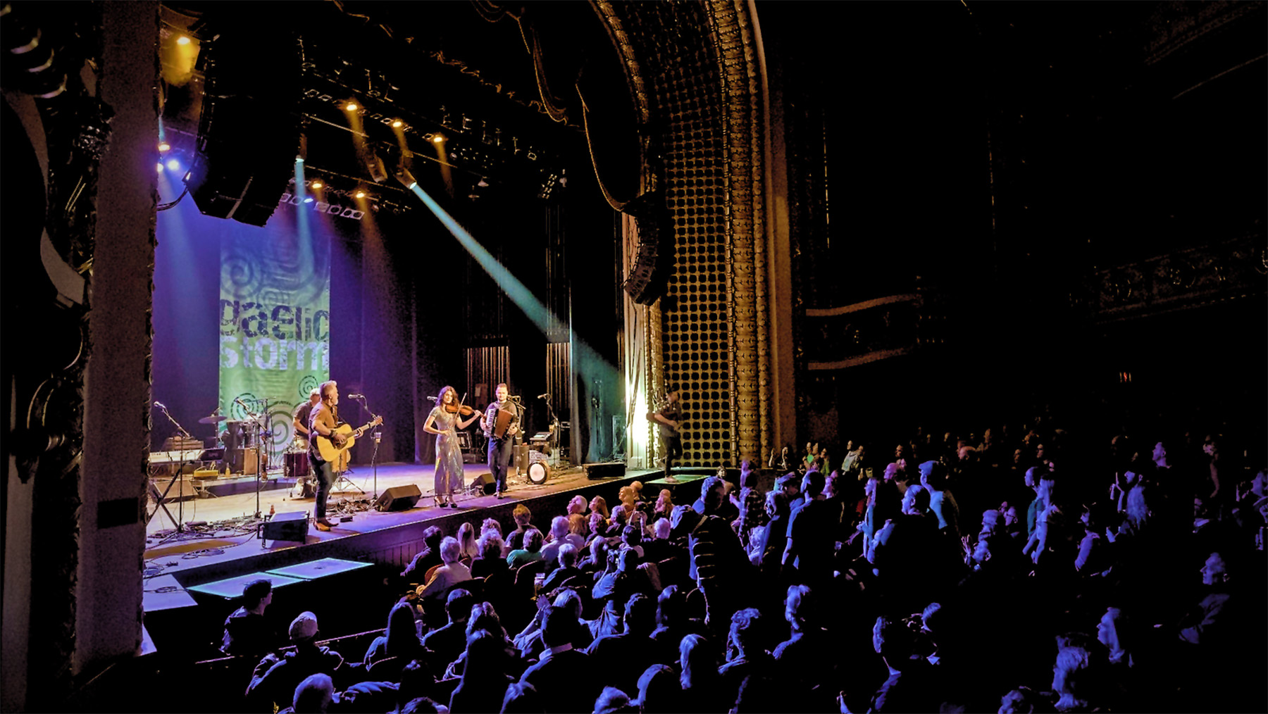A live concert scene with a band performing on stage, illuminated by stage lights, in front of an audience seated in a dark theater.