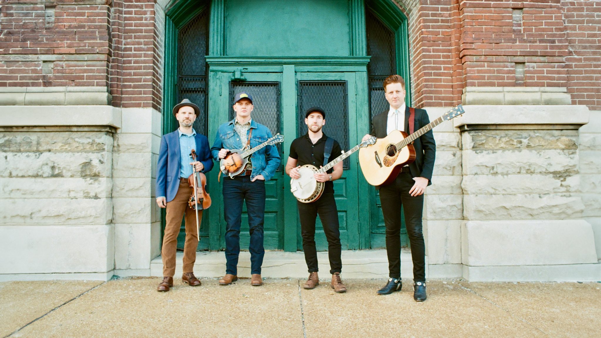 Four men stand in front of a large, green double door set in a brick and stone building. They are holding musical instruments including a violin, mandolin, banjo, and guitar. They are dressed in casual to semi-formal attire, including hats and jackets.