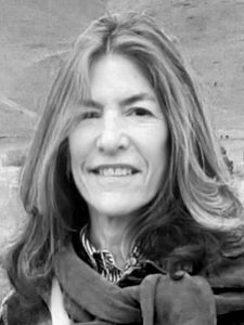 Amy Arnold Black and white photograph of a woman Amy Arnold with long hair, smiling at the camera. She is wearing a scarf and a patterned shirt, standing outdoors with a hill in the background. Her hair is parted in the middle and has waves.
